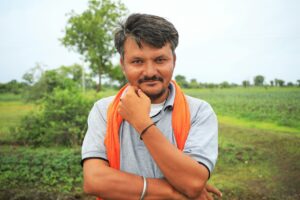 South Asian man with beard smiling confidently in a lush rural landscape, wearing an orange scarf.